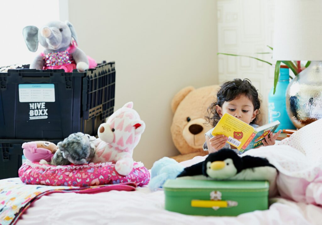 Image showing a girl on a bed reading a children's book, surrounded by stuffed toys